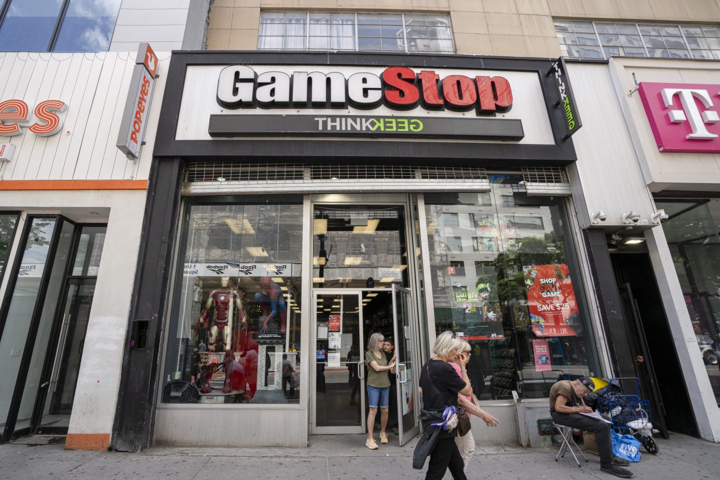 A GameStop in New York is seen from the sidewalk. People are seen passing the store and a young child is seen opening the door to leave the store.