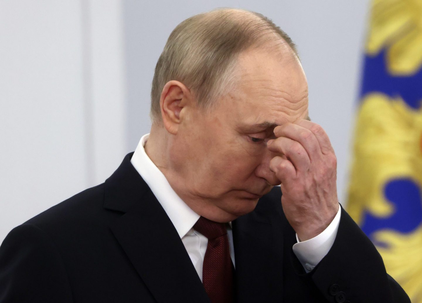 Russian President Vladimir Putin is captured in a close-up image, appearing deep in thought with his hand touching his forehead. He is wearing a black suit and a red tie, with a somber expression on his face.