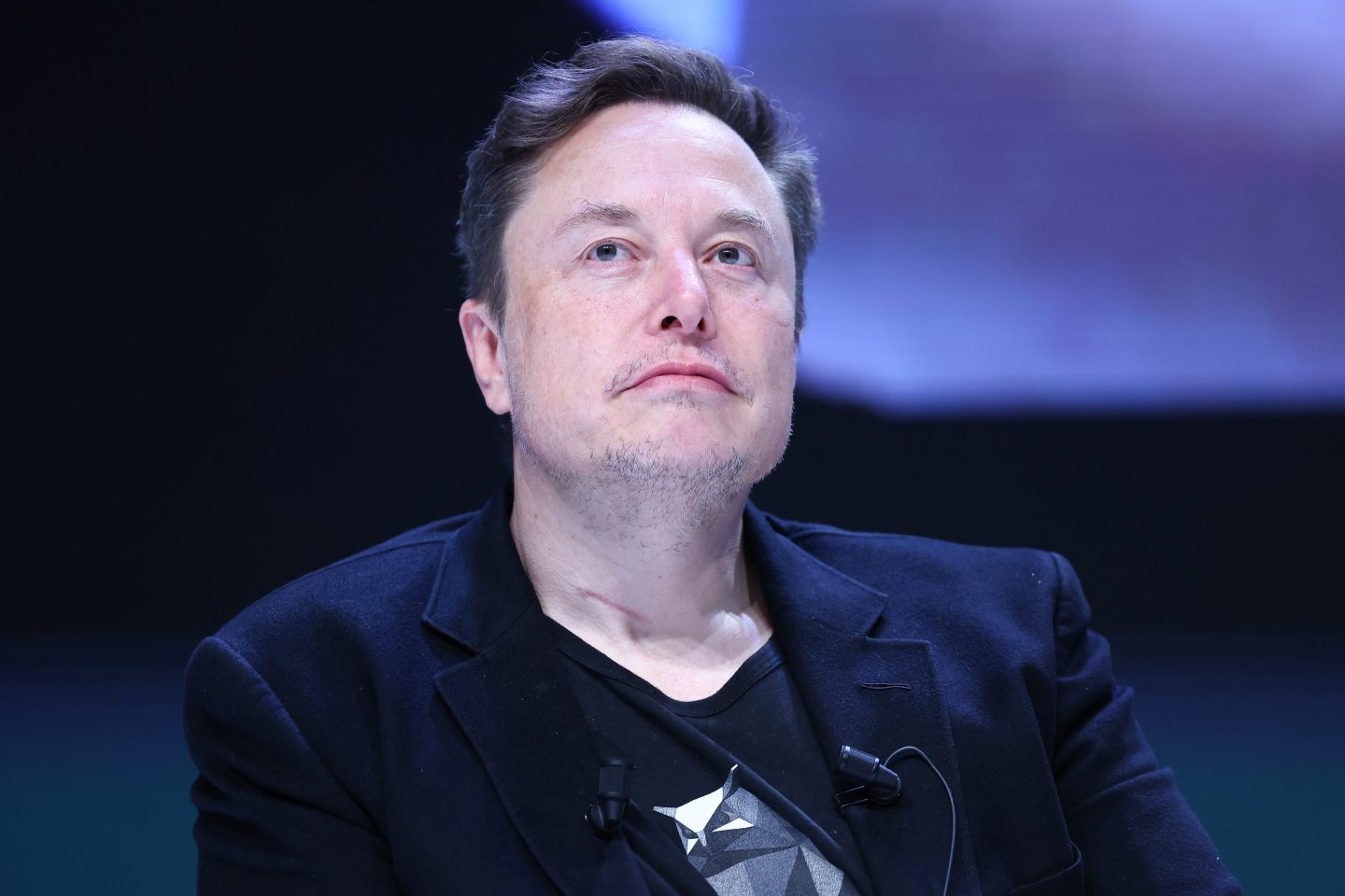 Elon Musk is seen at a panel event. He is wearing a tee shirt under a blazer and is looking up and out toward the audience.