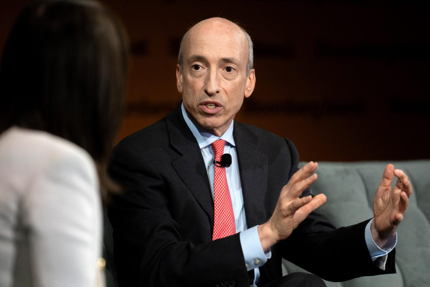 Gary Gensler, chairman of the US Securities and Exchange Commission (SEC), is seen at a panel speaking from a stage with a mic attached to his tie. He is mid-speech and gesturing with his hands. He wears a dark black suit with a light blue dress shirt and light red tie. In the foreground the host of the panel is blurred.