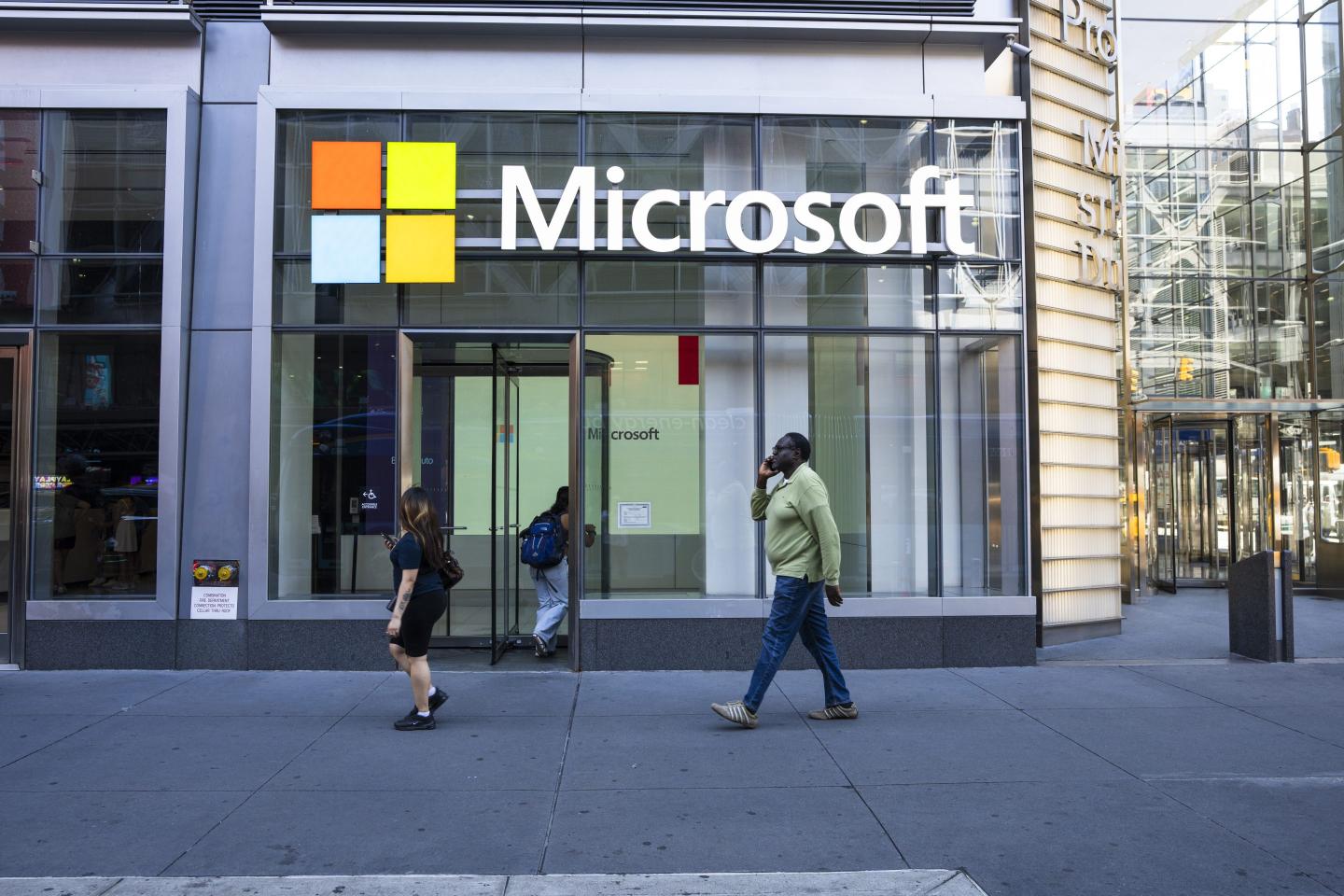 The image shows the exterior of a Microsoft store with the company's logo prominently displayed on the building. The logo features the iconic four-colored square next to the word "Microsoft" in white letters. Two people are walking by the storefront—one woman and one man, the latter talking on his phone. The scene captures a typical city sidewalk with glass walls and modern architecture surrounding the store.