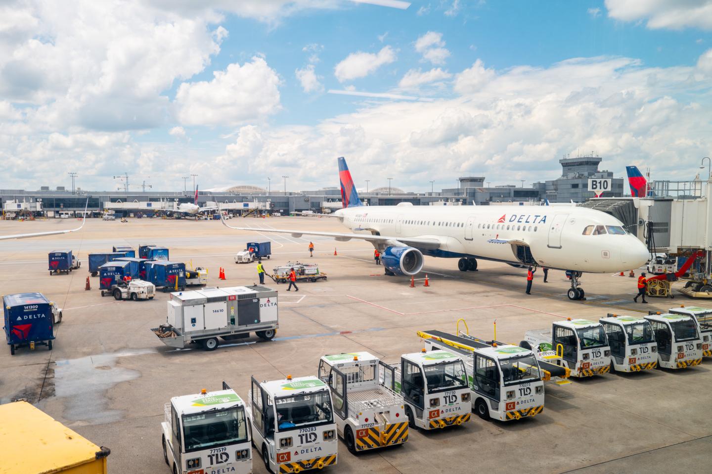 Delta Airlines aircraft parked at a busy airport gate, with ground crew working to load luggage and prepare the plane for departure. Several baggage carts and ground service vehicles are visible on the tarmac under a bright, partly cloudy sky.