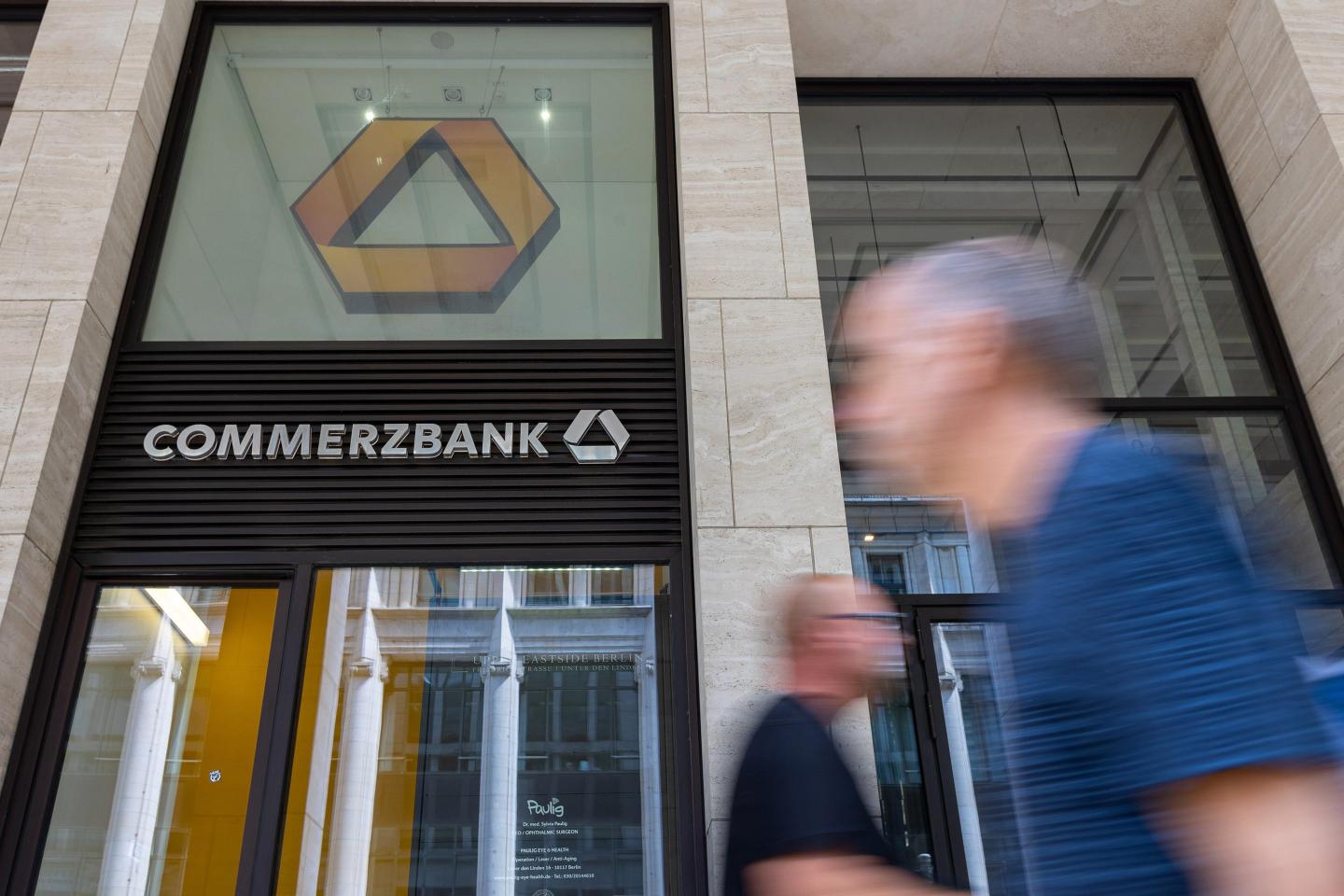 Blurred passersby walk in front of the Commerzbank logo outside an office building