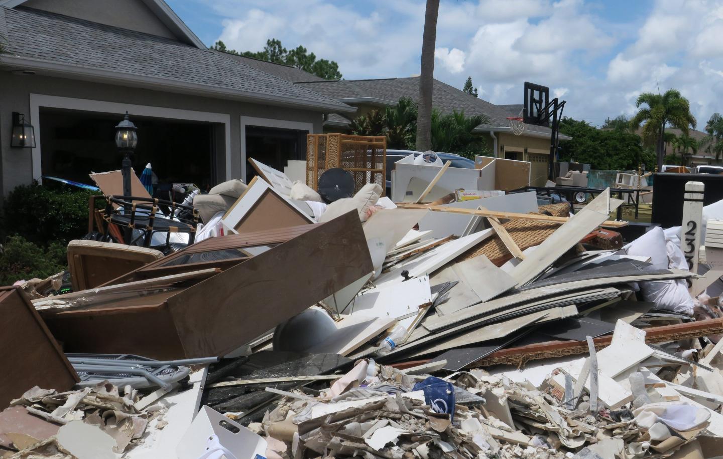 A large pile of debris and damaged household items is stacked in front of a suburban home, with the garage door open and more items visible inside. The pile includes broken furniture, drywall, and other household materials, the result of Tropical Storm Debby in Florida. The sky is clear with some clouds, and palm trees are visible in the background.