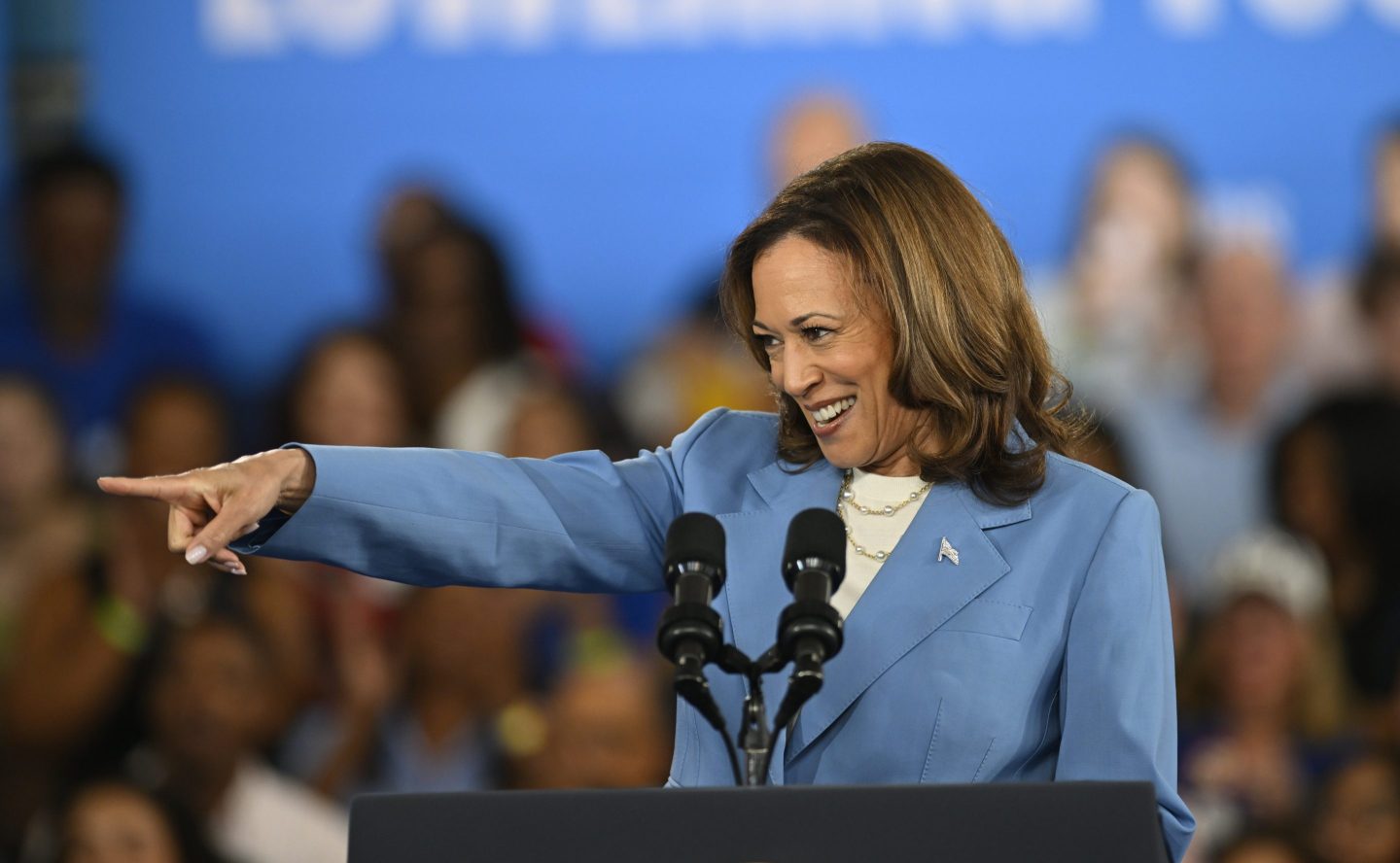 U.S. Vice President Kamala Harris, dressed in a light blue blazer, smiles and points enthusiastically while speaking at a podium with two microphones. The background features a blurred crowd, emphasizing the focus on Harris as she interacts with those in attendance.