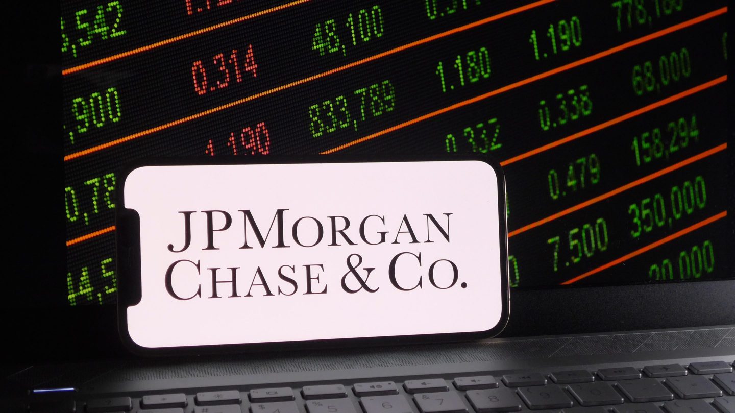 PMorgan Chase & Co. logo is displayed on a smartphone with a stock market and a laptop keyboard background.