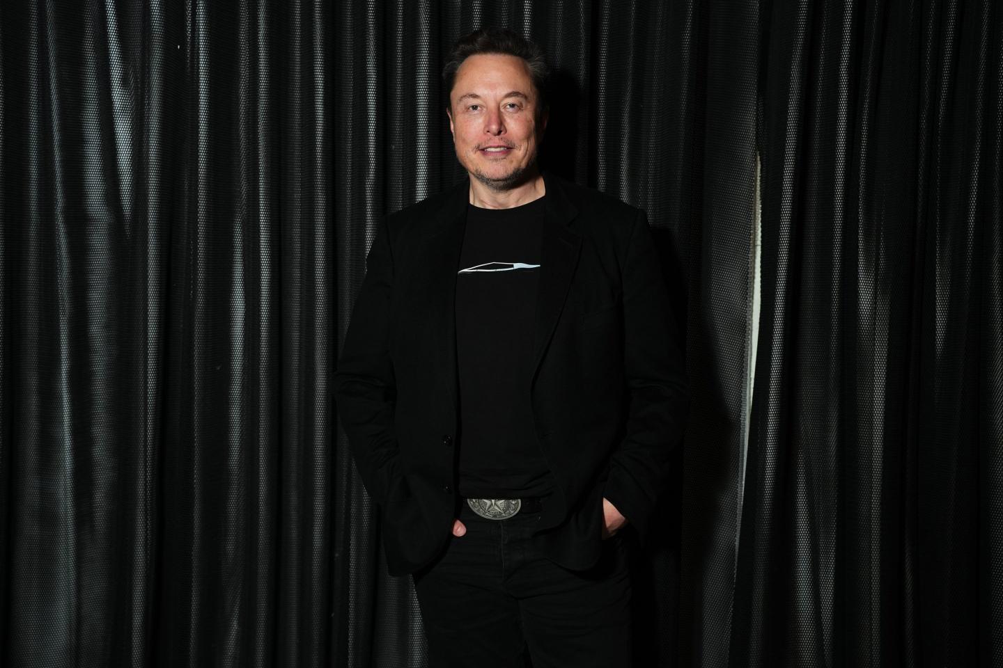 Elon Musk poses for a photo, standing in all black and smiling.
