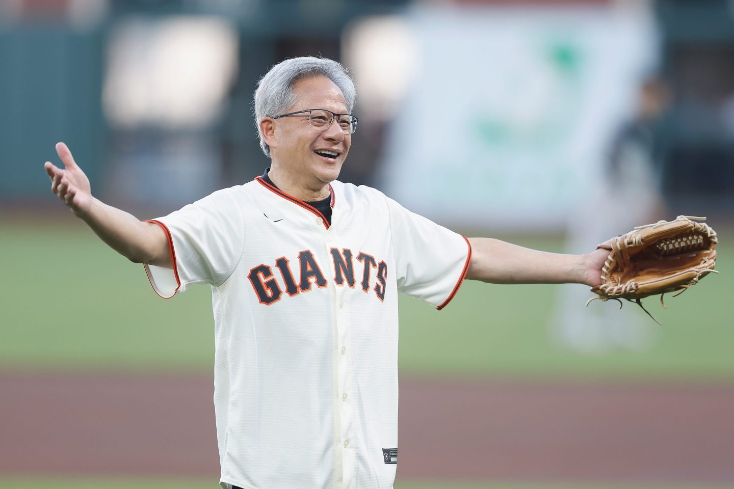 Jensen Huang, wearing a Giants baseball jersey and baseball glove on his left hand, stands with his arms out wide after apparently throwing out the first pitch.
