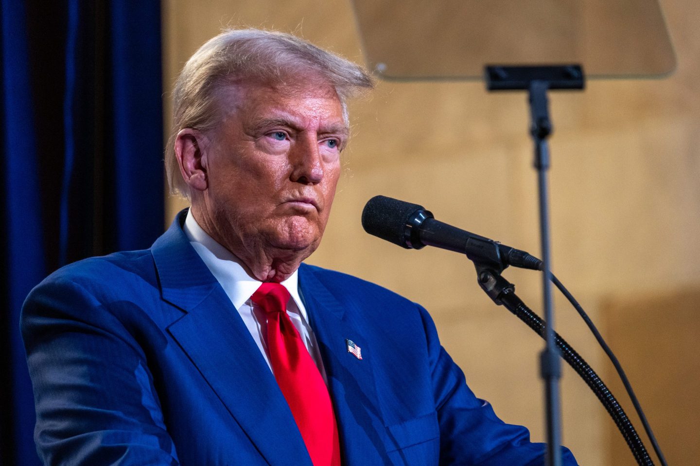 Former President Donald Trump stands at a mic with lips pursed He is wearing a blue blazer and a bright red tie with a white business shirt underneath.