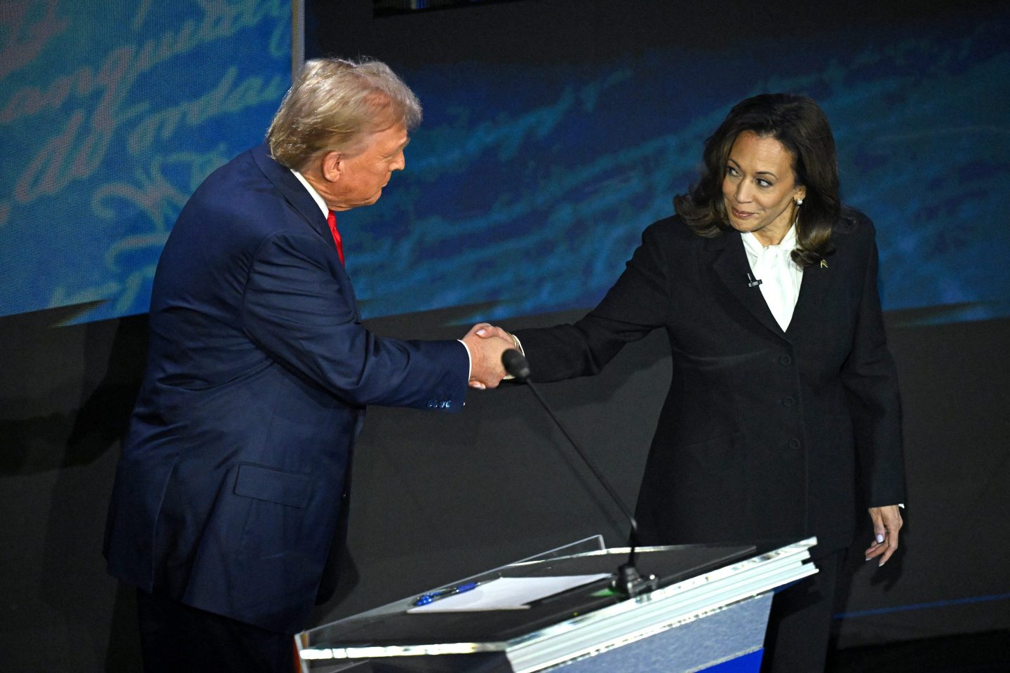 Former President Trump and Vice President Harris shake hands on the debate stage near Trump's podium. Both are wearing dark suits.