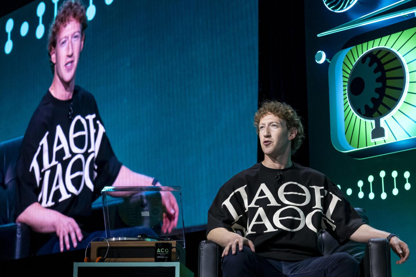 Mark Zuckerberg sits in a chair in front of a projection of himself on a screen above him.