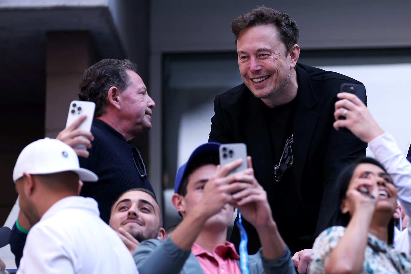 Elon Musk smiles as fans at the U.S. Open in the stands below him hold up their iPhones to take a selfie.
