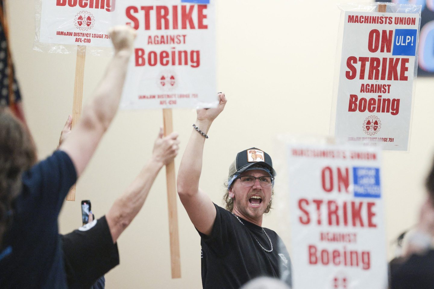 A man in the center of the image raises his right hand in a fist and shouts as fellow union members do the same or hold up signs calling for a strike against Boeing