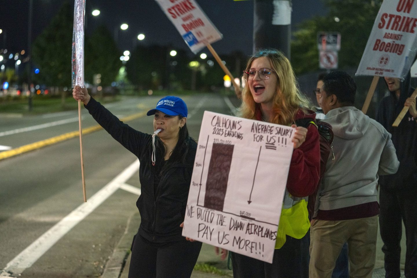 A woman with a sign comparing the average salary of Boeing's CEO to her and other factory employees chants at the side of the road with other strikers, including a woman next to her with a whistle and a blue hat that reads "Make Boeing Great Again."
