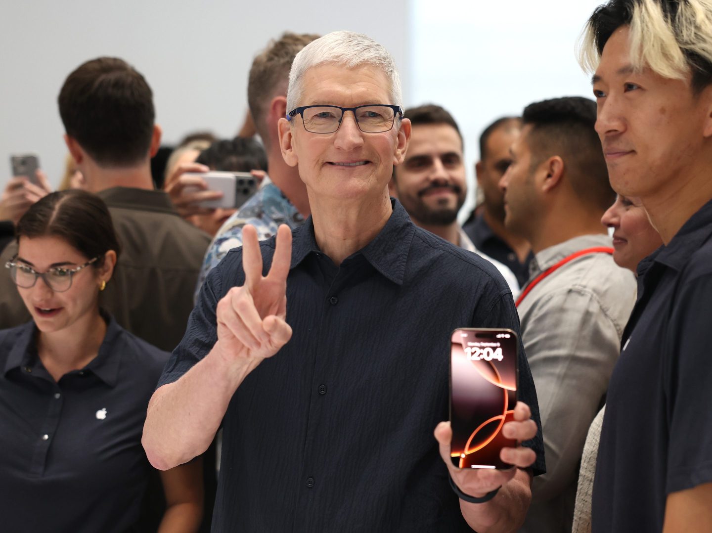 Apple CEO TIm Cook flashing a "V for Victory" sign on one hand while holding an iphone 16 in the other hand.