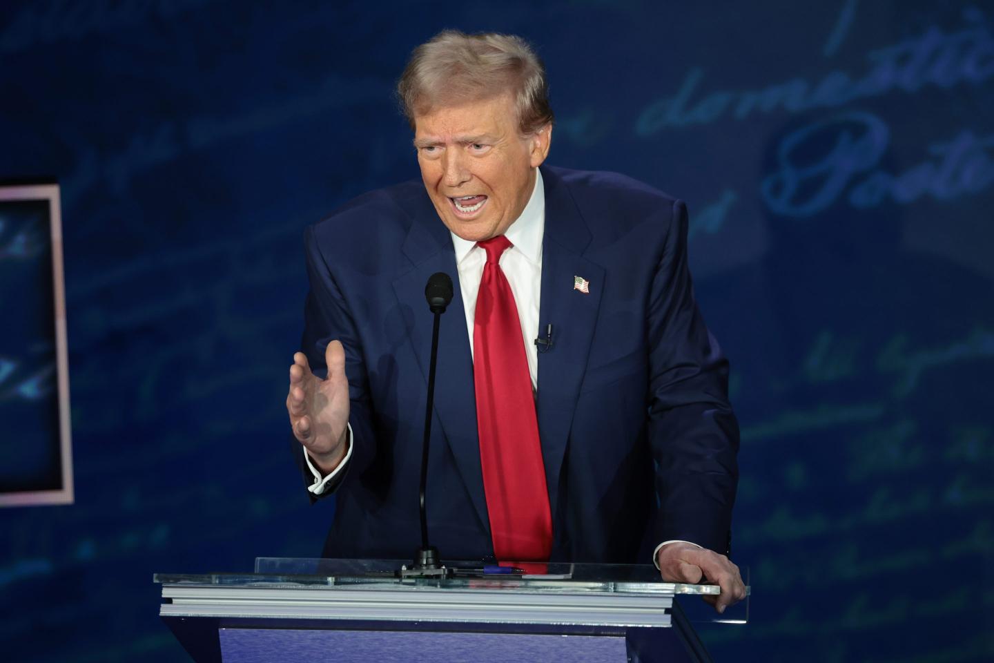 Former President Trump is seen behind the podium at his debate with Vice President Kamala Harris. he is wearing a dark navy suit with a long, bright red tie. He is gesturing with his right hand and his mouth is open as he speaks.