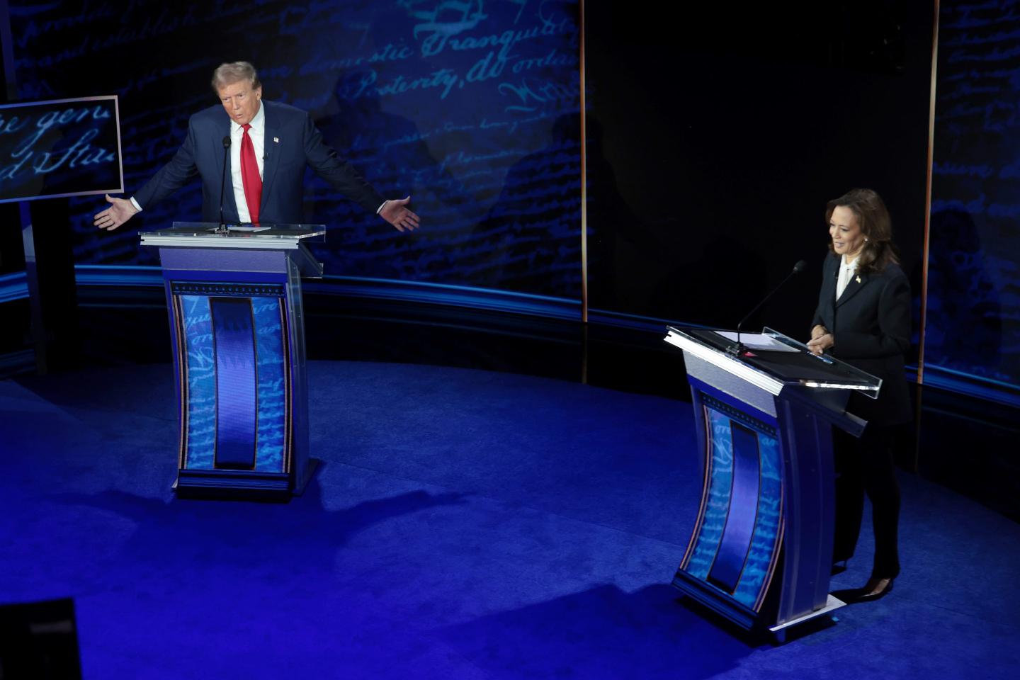 The image shows Donald Trump and Kamala Harris on stage during a debate. Trump, on the left, is standing behind a podium, gesturing with his arms spread wide as he speaks. He is wearing a navy suit with a red tie. Harris, on the right, is smiling and looking down at her podium, possibly listening or preparing to respond. She is dressed in a dark suit with a white blouse. The stage has a deep blue color, with script-like text in the background adding to the formal debate setting. The podiums are sleek with blue panels, and the lighting creates distinct shadows on the stage floor.
