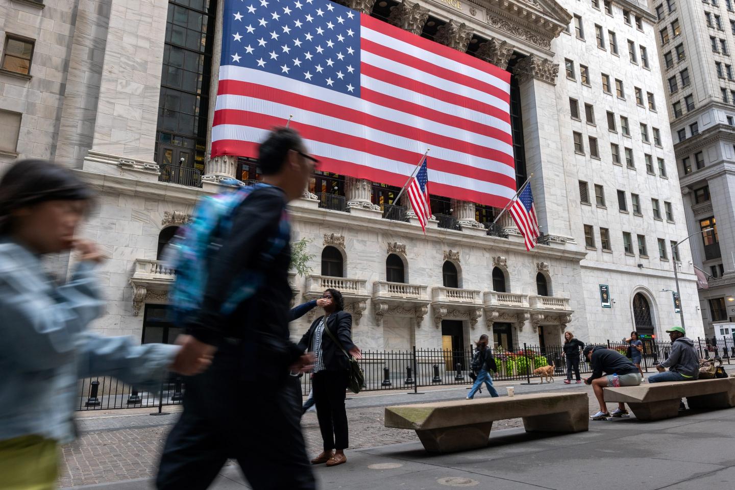 The image shows pedestrians walking in front of the New York Stock Exchange building, which features a large American flag draped across its front facade. The building is adorned with classic architecture, including columns, and two smaller American flags flanking the main entrance. Some people are walking by, blurred in motion, while others sit on nearby benches. The scene captures the bustling nature of Wall Street, set against the backdrop of one of the most iconic financial institutions in the world.