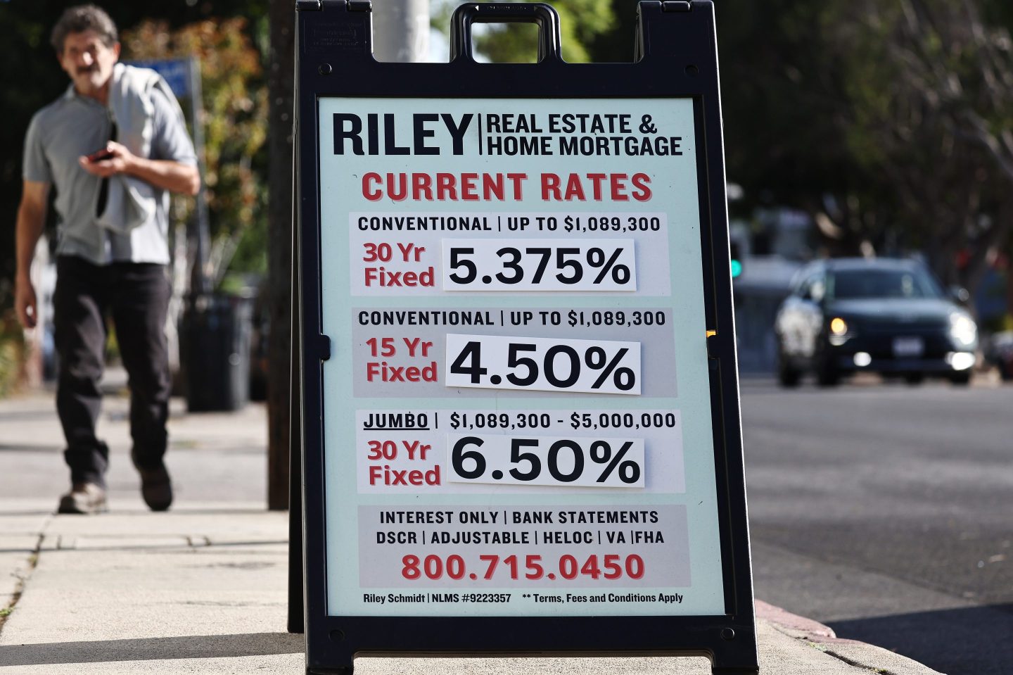 A street sign for "Riley Real Estate & Home Mortgage" displays current mortgage rates. The rates listed are 5.375% for a 30-year fixed conventional loan up to $1,089,300, 4.50% for a 15-year fixed conventional loan up to $1,089,300, and 6.50% for a 30-year fixed jumbo loan ranging from $1,089,300 to $5,000,000. The sign also includes information about other services such as interest-only loans, bank statement loans, DSCR loans, adjustable-rate mortgages, HELOC, VA loans, and FHA loans, with a contact phone number at the bottom.