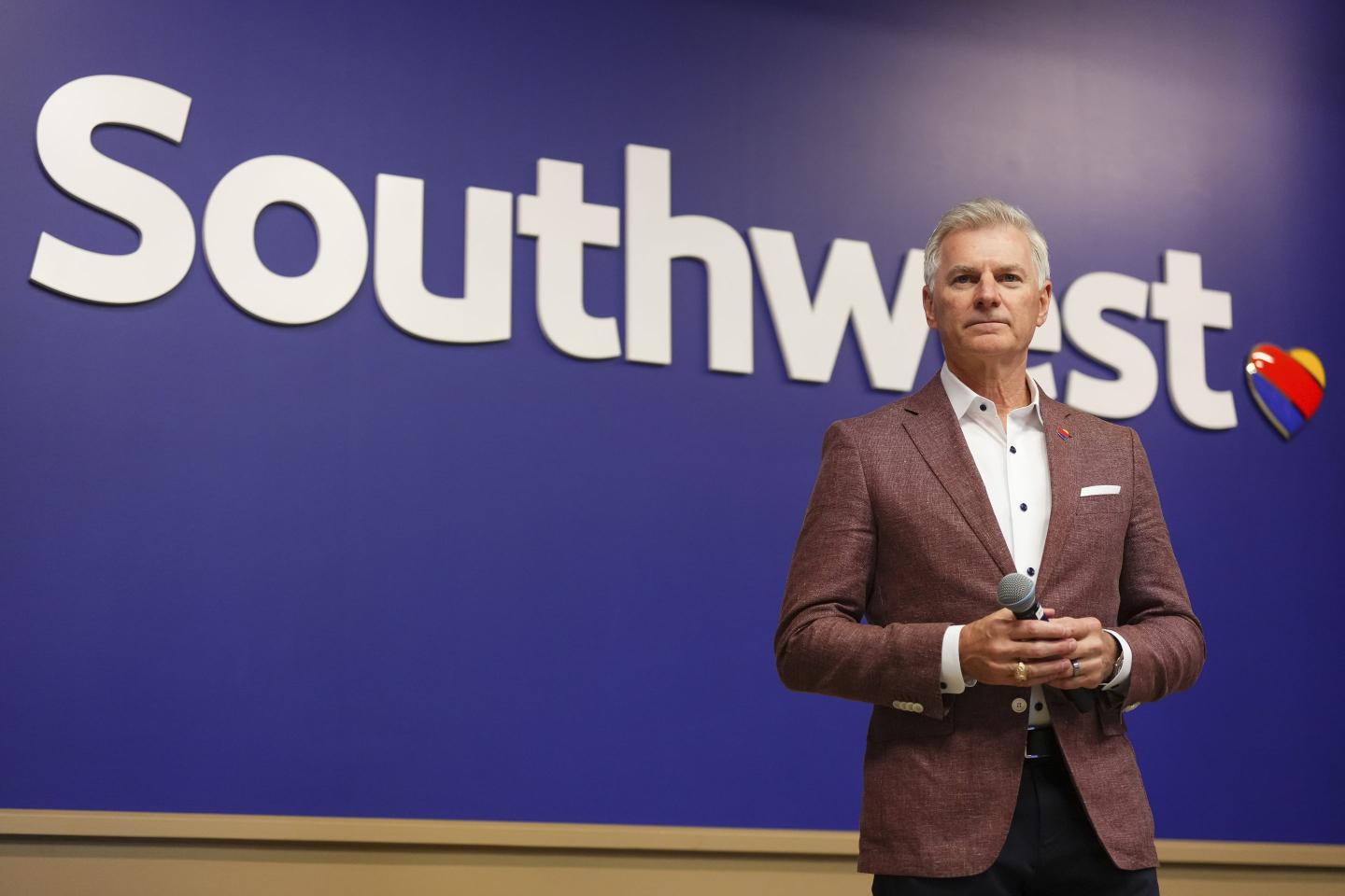 Southwest CEO Bob Jordan, wearing a white button-down shirt and reddish-brown jacket, holds a microphone in front of a wall with the Southwest logo.