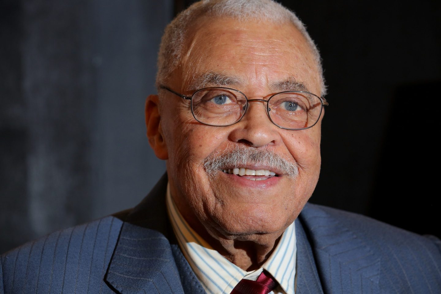 Actor James Earl Jones is seen smiling at the premier of an event. He is wearing glasses and a navy coat blazer with a red tie.