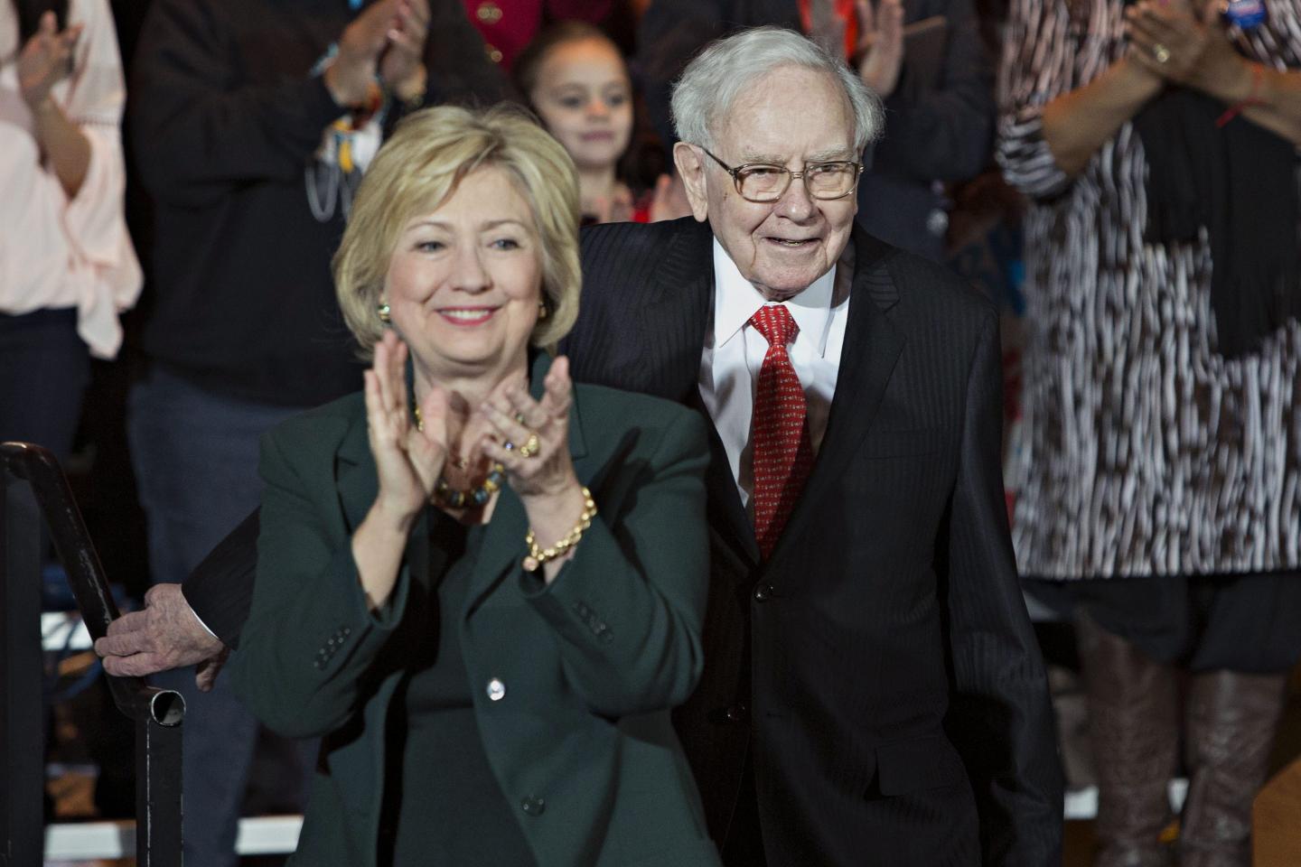 Hillary Clinton, dressed in a green jacket, claps as Warren Buffett, dressed in a black suit, white shirt and red tie, walks behind.