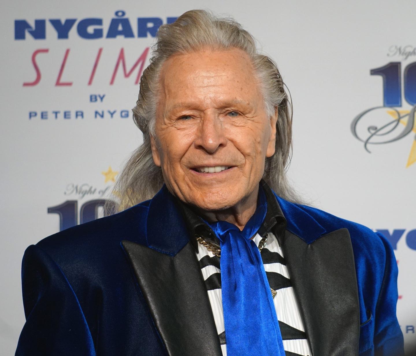 Peter Nygard poses for press at a red carpet event. He is wearing a dark blue and black coat with a brighter, royal blue tie. His shirt has a black and white pattern beneath the coat.