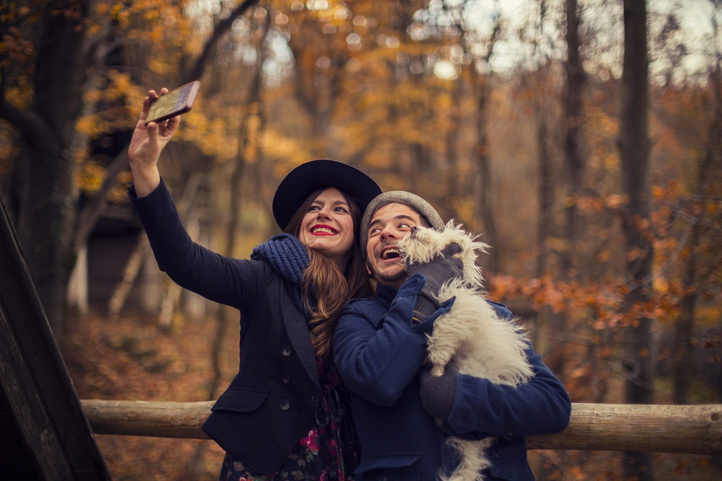 A man and woman pose with a small dog and take a selfie in front of autumn foliage.