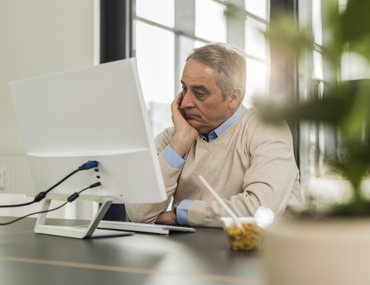 An older man sits in front of a computer with his head in his hand.