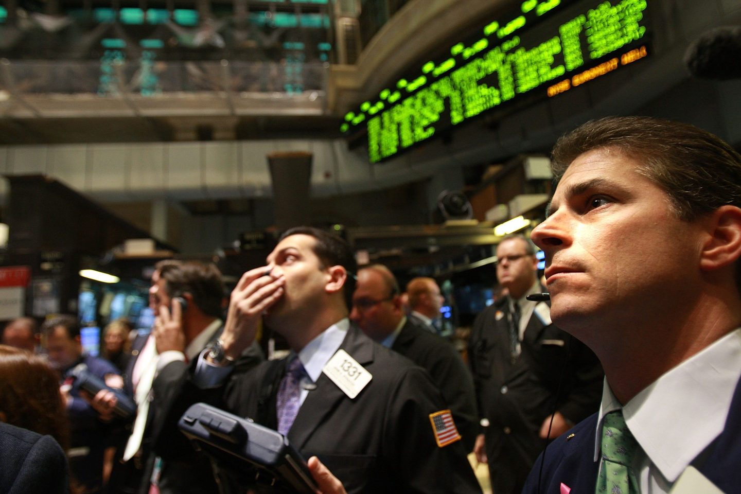 One man in center of the photo on the floor of the New York Stock Exchange covers one hand over his mouth (his other hand is holding a tablet) while another man in the foreground looks up in the same direction with a worried look on his face.