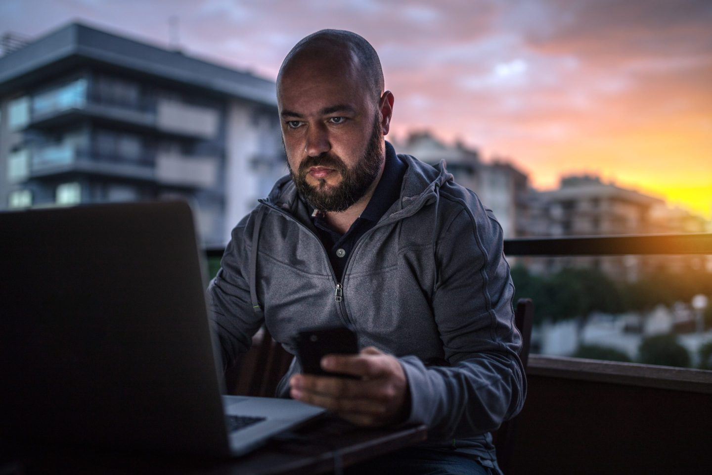 Bald and bearded men uses laptop and smartphone at sunset