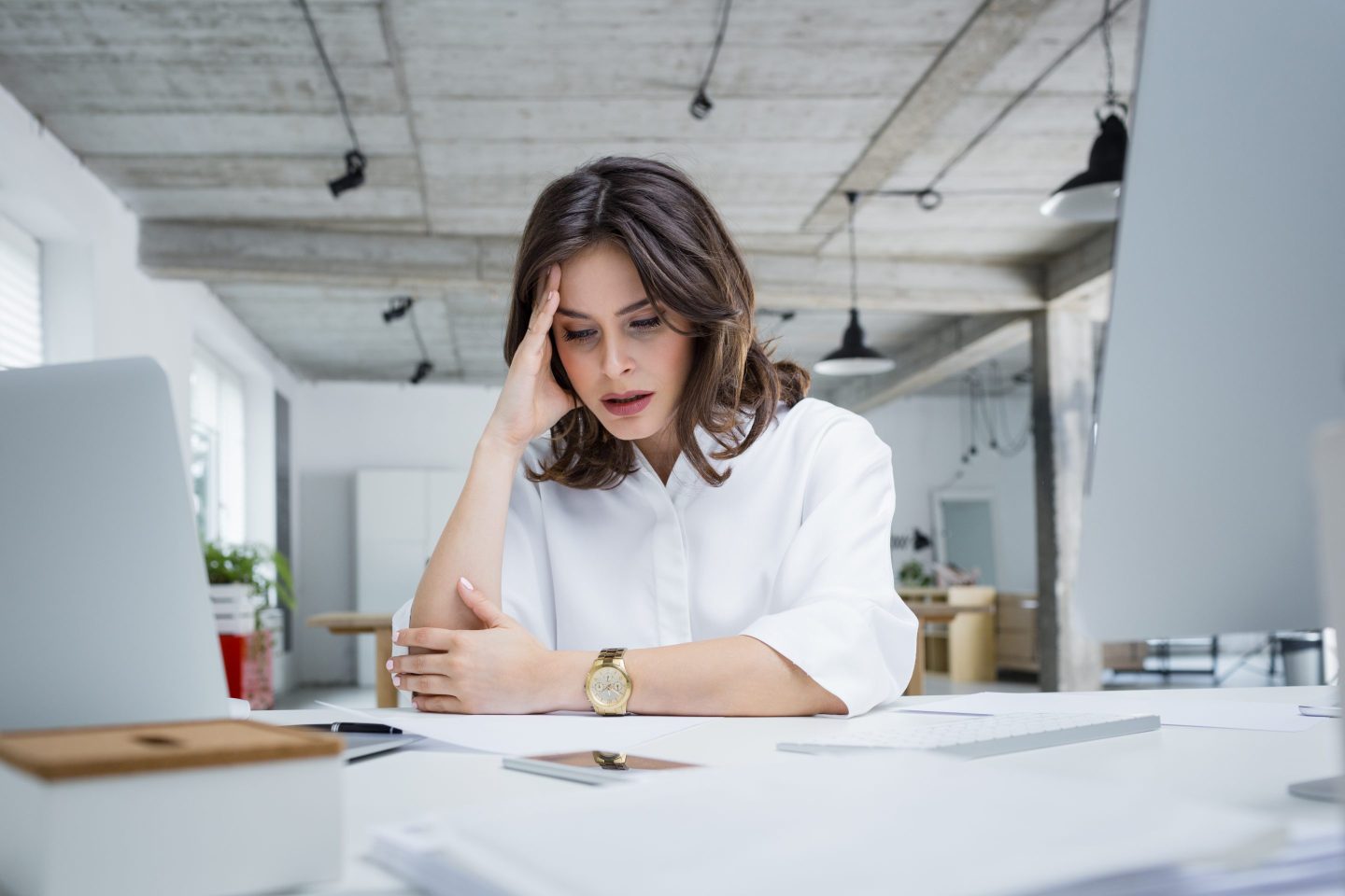 A white woman with brown hair holds her head as she looks at papers on her desk.