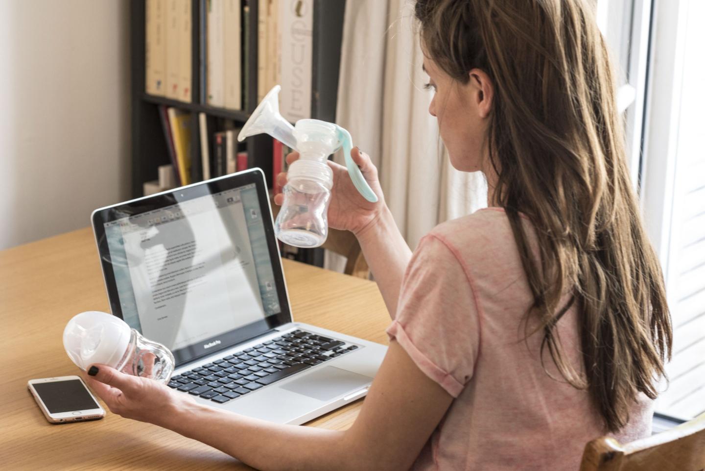 A woman is sitting at a desk in front of an open laptop, holding a manual breast pump in one hand and a baby bottle in the other. The image captures the balance of multitasking between professional duties and motherhood, highlighting the challenges faced by working moms who pump breast milk.