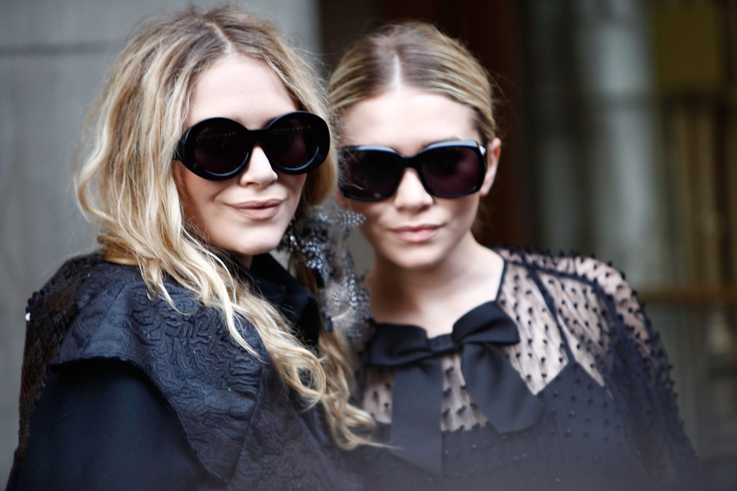 Mary-Kate and Ashley Olsen appear wearing all black and large black sunglasses. They are looking at the camera smiling.