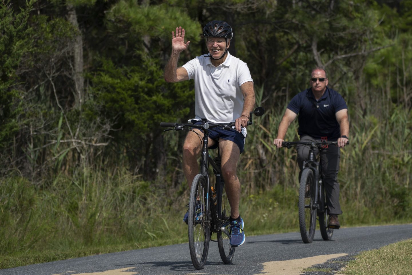 President Joe Biden waves to a cheering crowd as he rides his bike followed by a Secret Service agent at Gordons Pond in Rehoboth Beach, Del., on Aug. 11, 2024.