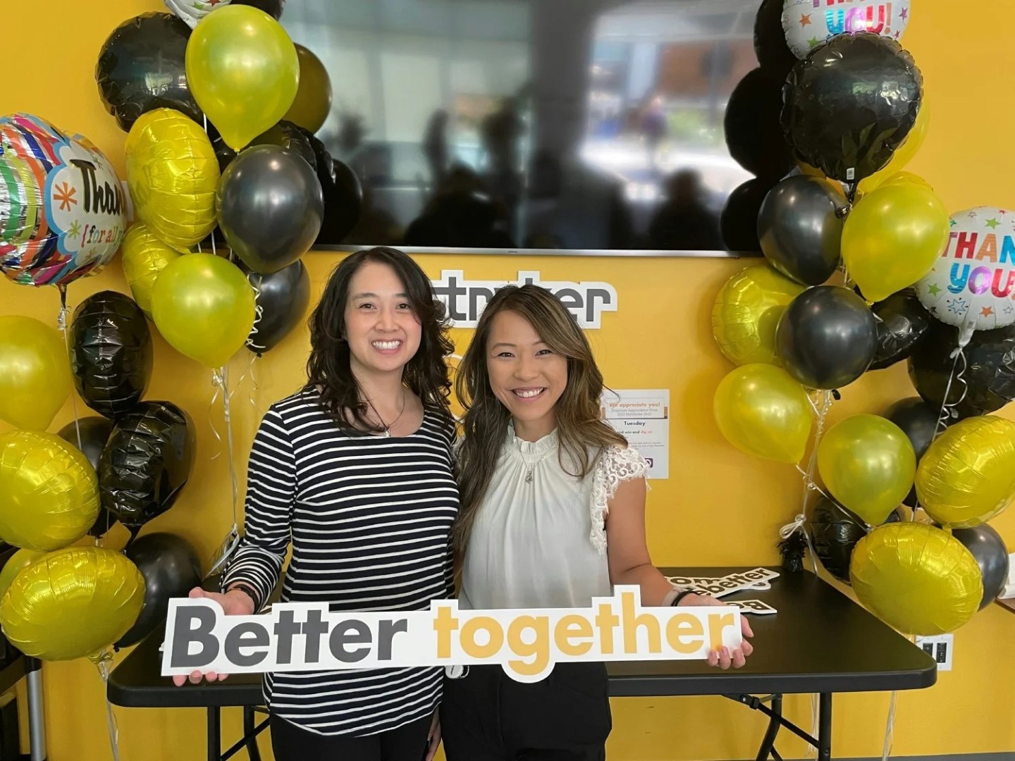 Two female Stryker employees stand in front of employees holding a "better together" sign.