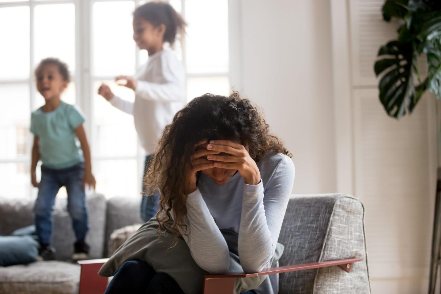 Black single frustrated woman hold her head with hands sitting on chair in living room, playful kids jumping on couch on a background.