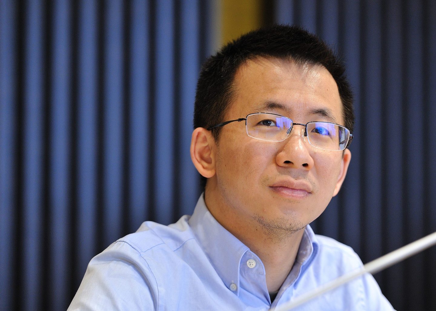 Zhang Yiming, wearing a blue Oxford shirt and glasses, looks pensive.