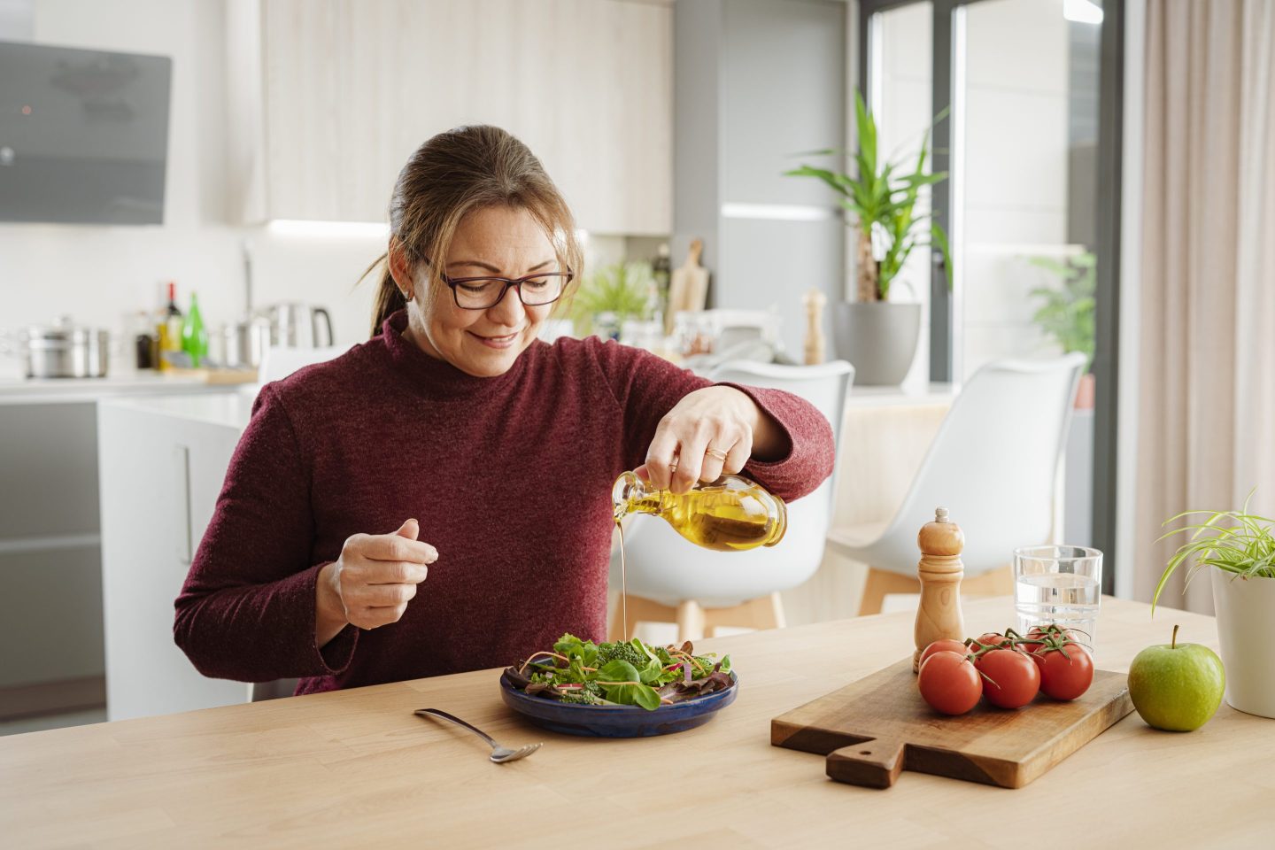 Portrait of a smiling woman dressing on healthy green salad with olive oil. She is sitting at dining table with modern kitchen at background.