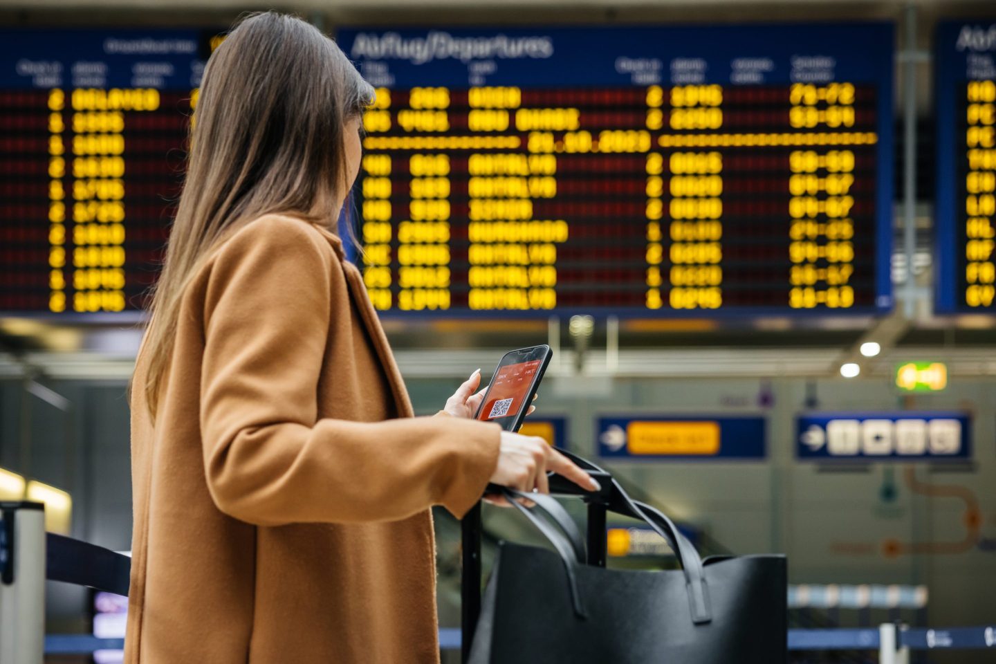 A woman traveler checks her mobile phone in front of the airport&#8217;s flight information board, preparing for her upcoming solo journey.