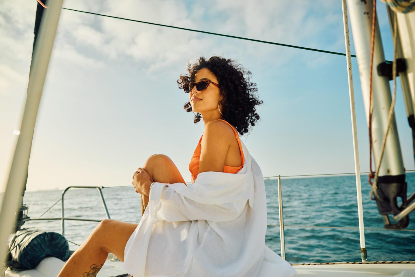 A contemplative young woman lounges on the deck of a sailing yacht, draped in a casual white shirt over her vibrant orange swimwear. Her denim shorts and stylish sunglasses speak to a laid-back day at sea, with her curly hair tousled by the gentle ocean breeze.