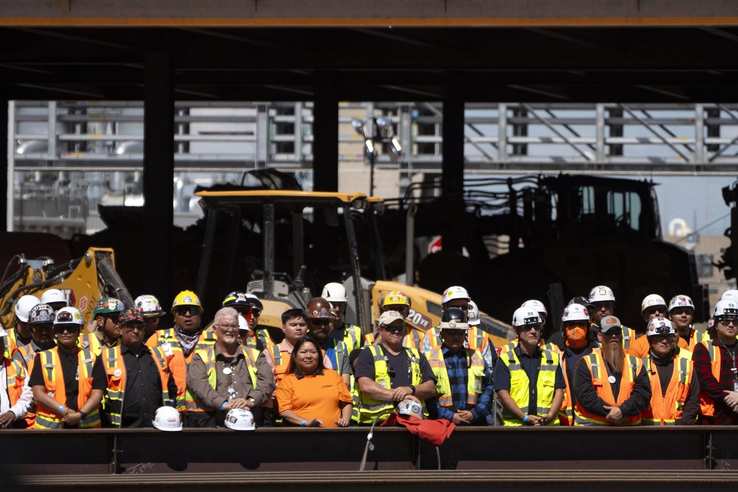 Men and women in construction gear watch a speech from President Biden behind a big steel beam and construction equipment in the background.
