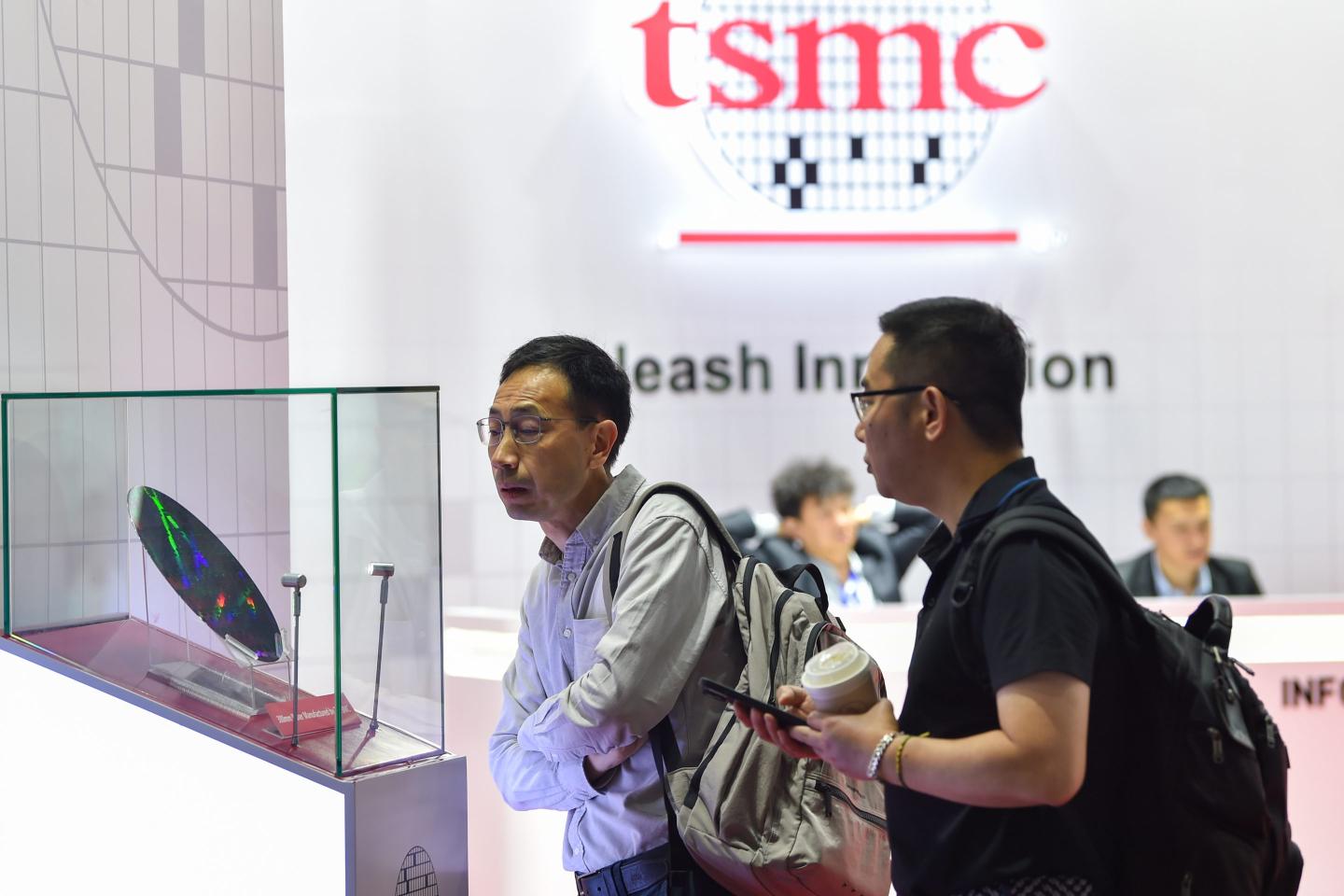 Two men peer at a massive AI chip held in a glass box. The logo of TSMC is visible in the background.