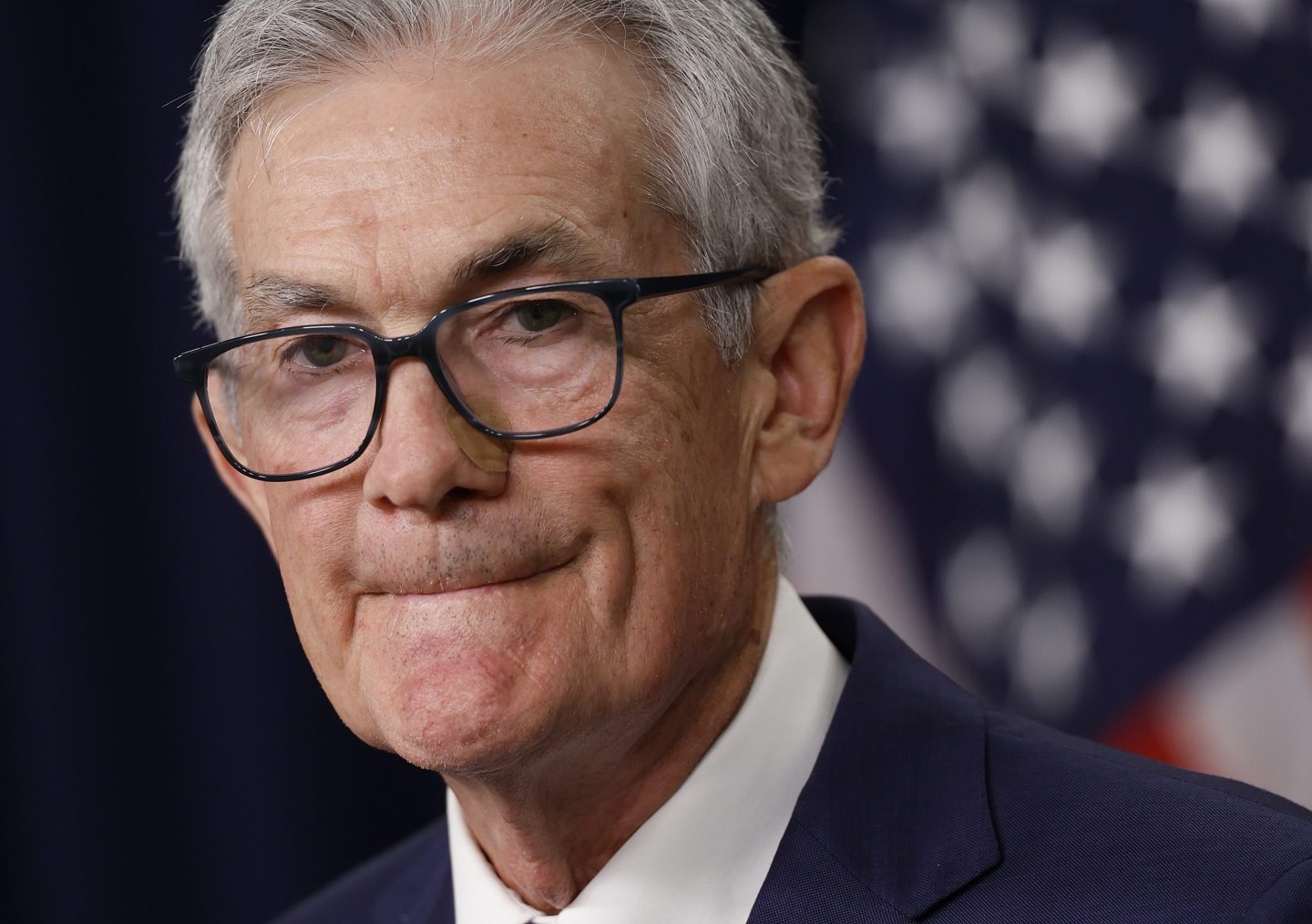 Federal Reserve Bank chair Jerome Powell is seen in front of the American flag, wearing a suit. His lips are tightly shut.