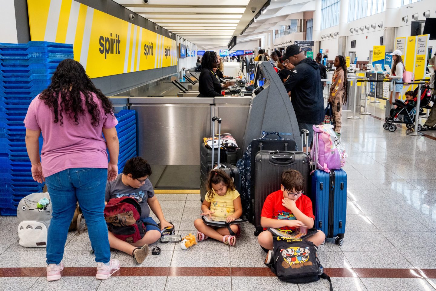 Children sit with backpacks, books, and suitcases as a woman talks with them near check-in desks for Spirit Airlines.