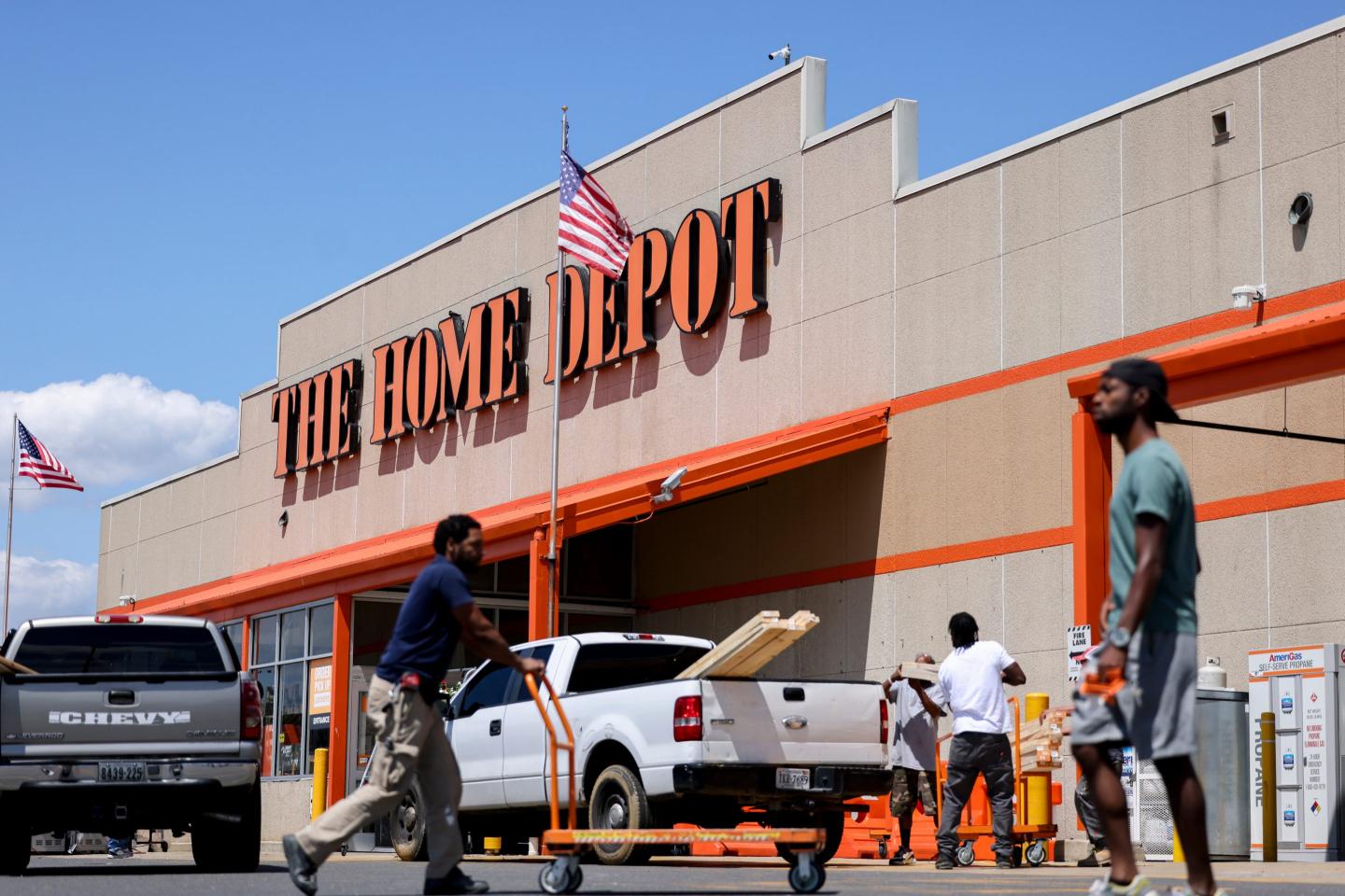 A Home Depot store is seen from the parking lot, with people entering the store and a white truck loading lumber.