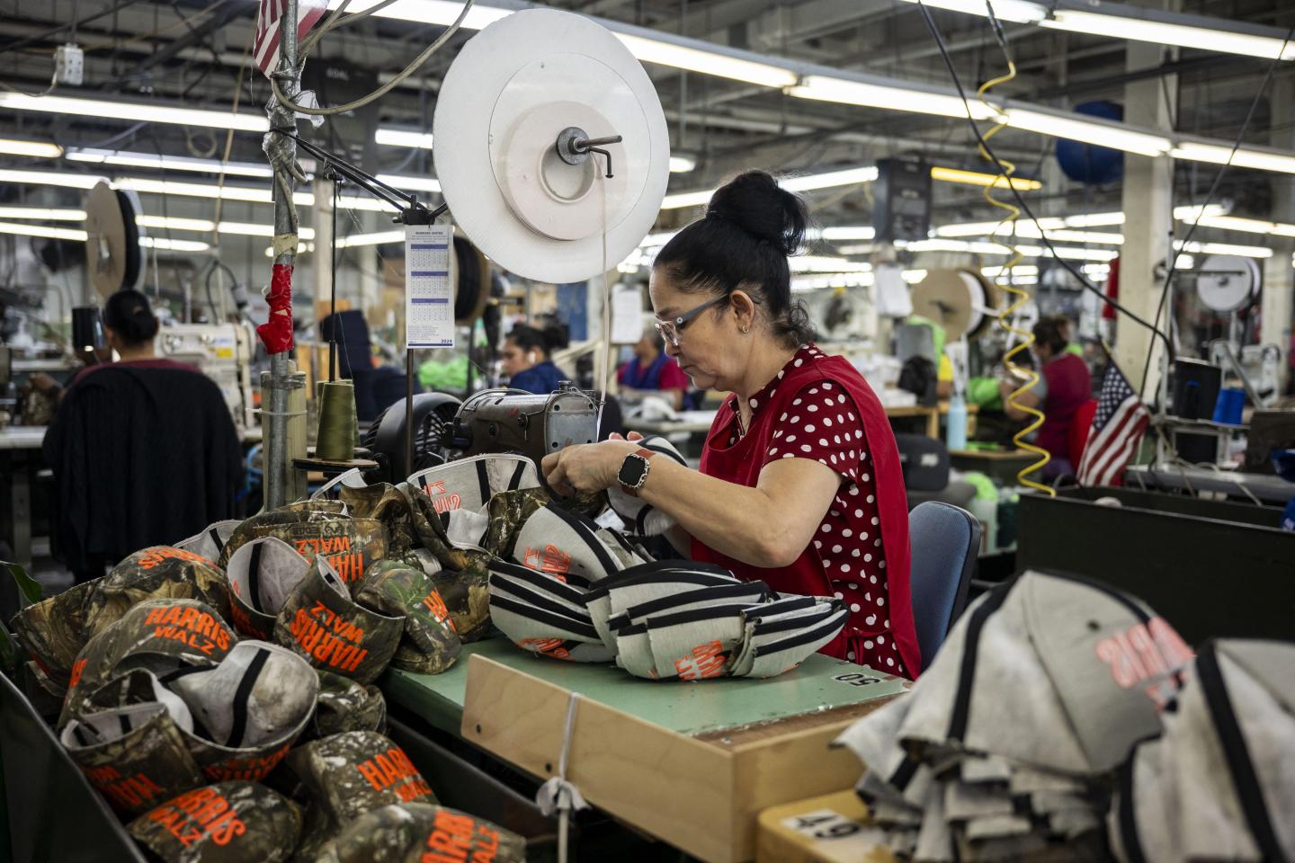 A woman in an apron works through stitching together a pile of camo hats.