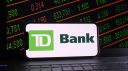 In this photo illustration, Toronto-Dominion Bank logo is displayed on a smartphone with a stock market and a laptop keyboard background.