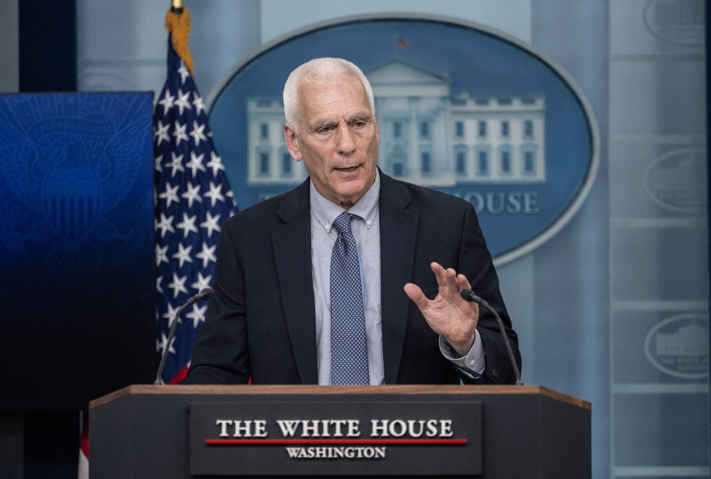 Jared Bernstein stands at a podium and raises his left hand as he speaks at a press briefing.
