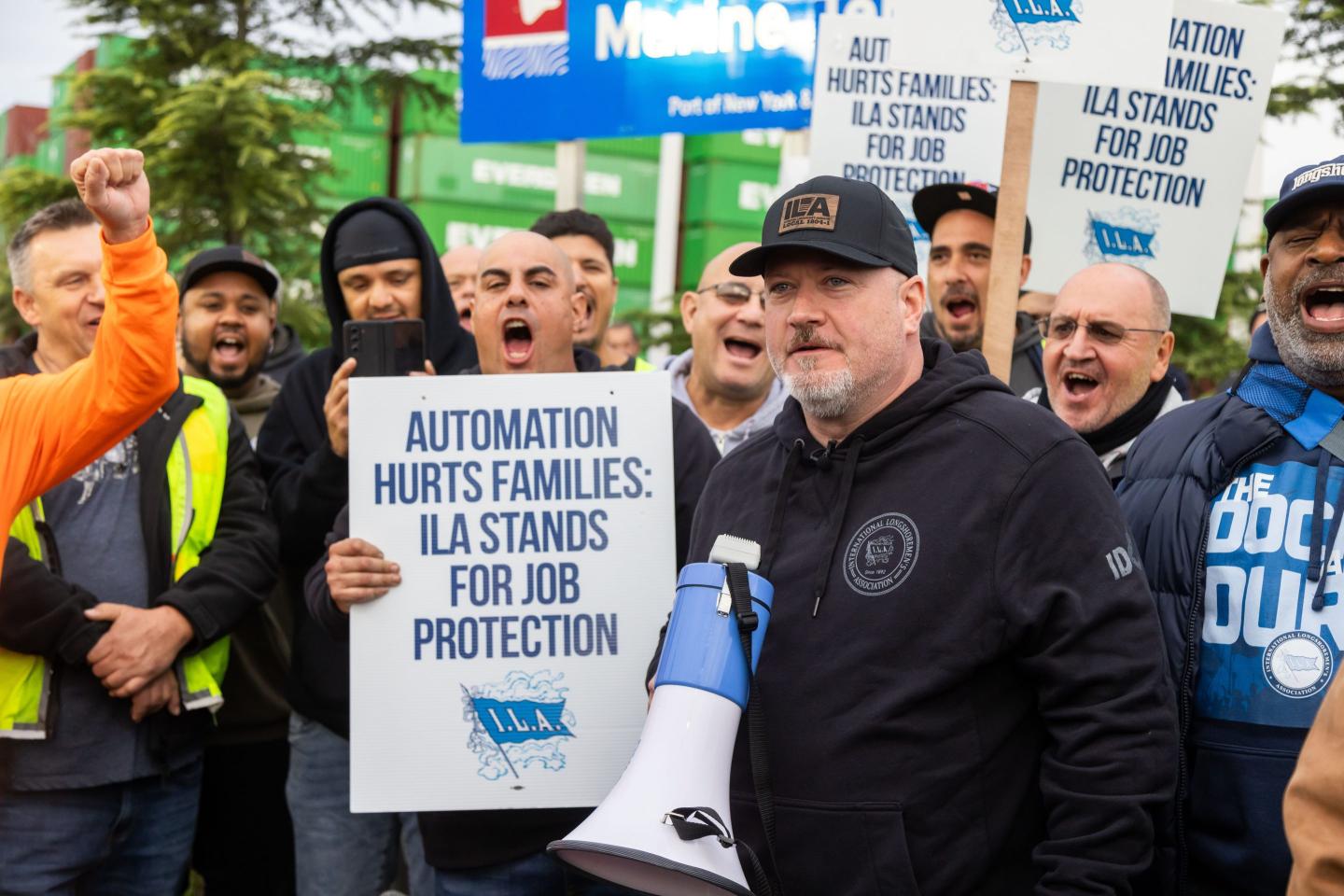 Striking longshoremen carry signs and cheer behind a man speaking to them with a megaphone.